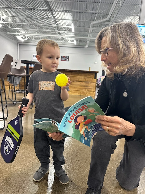Young toddler boy holding a pickleball paddle and yellow ball next to Karen Worthy holding Bend Your Knees Louise Pickleball Book